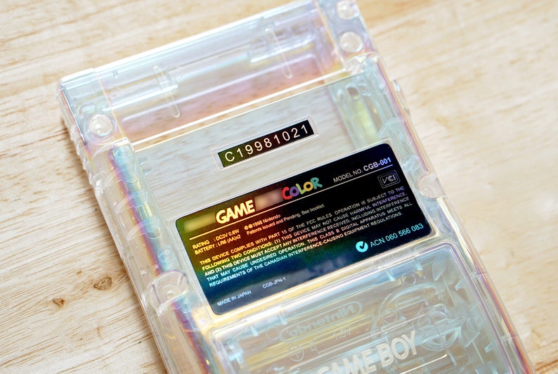 May include: A clear Game Boy Color handheld gaming console with a gold label that reads "GAME COLOR" and includes the model number "CGB-001". The label also includes the rating, battery, and other information.