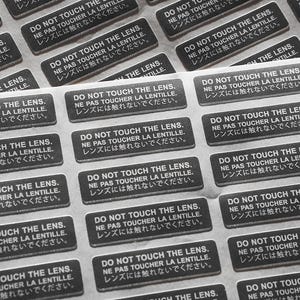 May include: A sheet of stickers with the text "Do Not Touch The Lens. Ne Pas Toucher La Lentille. レンズには触れないでください." repeated multiple times in black text on a white background.