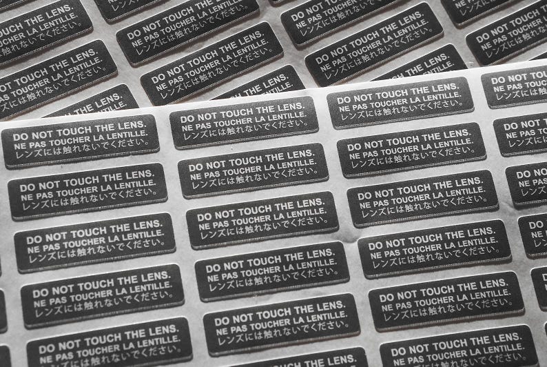 May include: A sheet of stickers with the text "Do Not Touch The Lens. Ne Pas Toucher La Lentille. レンズには触れないでください." repeated multiple times in black text on a white background.