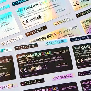 May include: A close-up of several holographic stickers for a Game Boy Color handheld video game console. The stickers are black, silver, and blue with white text. The text on the stickers includes "Game Boy Color", "Model No. CGB-001", "Rating: DC3V 0.8W", "Battery: LR6 (AA)x2 or Adapter Input: DC3V 300mA", "1998 Nintendo Patents Issued and Pending, See Booklet", "CE", "LGAD", "Made in Japan", and "CGB-EUR".