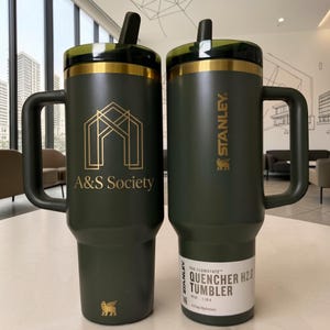 Könnte beinhalten: Zwei dunkelolivgrüne 1,18-Liter-Stanley-Quencher-H2.0-Becher mit Griffen und dunklen Deckeln. Der linke Becher zeigt ein goldenes geometrisches Logo und den Text A&S Society. Der rechte Becher trägt das Stanley-Logo und ein Produktetikett.