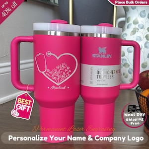 May include: Two pink Stanley Quencher H2.0 tumblers with white lids and handles. One features a heart design with a stethoscope and floral elements, the other has the Stanley logo. The tumblers are 1.18L and suitable for all-day hydration.