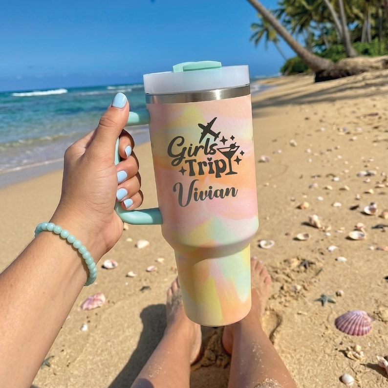 May include: A pastel tie-dye tumbler with a light blue handle and lid. The cup features black text that reads Girls Trip and Vivian, accompanied by a small airplane icon and a martini glass graphic, set against a beach and ocean background.
