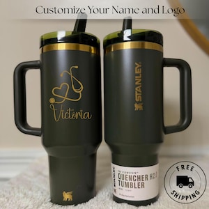 Peut inclure: Deux gobelets Stanley Quencher H2.0 vert foncé avec des poignées et des couvercles noirs. L'un a un motif de cœur et de stéthoscope doré avec le nom "Victoria". L'autre porte le logo Stanley. Les deux ont une paille.