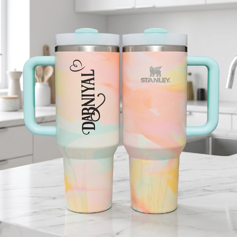 May include: Two stainless steel tumblers with handles and light blue lids, featuring a pastel watercolor pattern in shades of peach, yellow, and mint. One tumbler displays the name DABNIYAL in black script, while the other shows the Stanley logo.