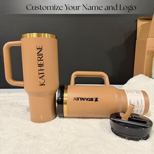 以下が含まれることがあります： 2つの断熱タンブラーと蓋が表示されています。1つのタンブラーにはハンドルがあり、「KATHERINE」という名前が印刷されています。もう1つのタンブラーは水平で、「STANLEY」というブランド名が印刷されています。どちらもタン色で、ゴールドのアクセントが付いています。上部のテキストは「名前とロゴをカスタマイズ」と書かれています。
