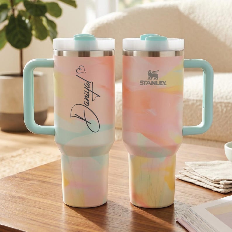 May include: Two tall insulated tumblers with handles and light blue lids, featuring a pastel watercolor pattern in pink, yellow, and blue. One tumbler displays the name Daniyal in black script with a heart, while the other features the Stanley logo.