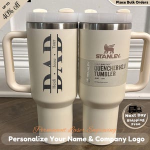 Peut inclure: Deux gobelets Stanley Quencher H2.0 crème avec poignées. L'un est gravé avec "DAD" et des noms, l'autre a une étiquette Stanley. Les deux ont des couvercles blancs et une capacité de 1,18 L. L'image comprend le texte : "Personnalisez votre nom et le logo de votre entreprise."