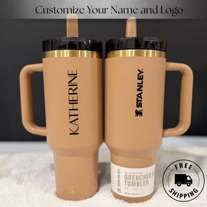 以下が含まれることがあります： 黒い蓋と金色のアクセントが付いた、タン色のスタンレー・クエンチャータンブラー2個。1つには「KATHERINE」という名前、もう1つには「STANLEY」のロゴが入っています。「Customize Your Name and Logo」というテキストが上部にあります。