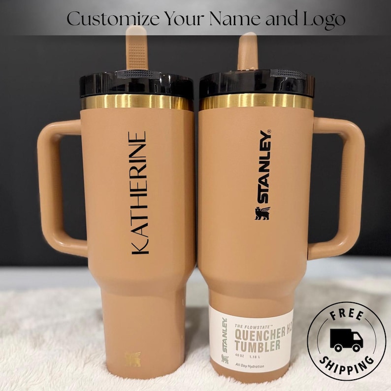以下が含まれることがあります： 黒い蓋と金色のアクセントが付いた、タン色のスタンレー・クエンチャータンブラー2個。1つには「KATHERINE」という名前、もう1つには「STANLEY」のロゴが入っています。「Customize Your Name and Logo」というテキストが上部にあります。