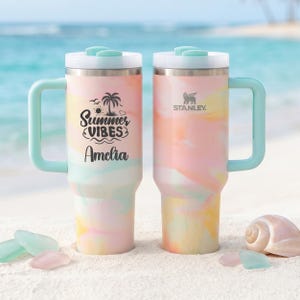 May include: Two stainless steel tumblers with handles and light blue lids, featuring a pastel pink, yellow, and blue marbled pattern. One tumbler displays the black text Summer Vibes and the name Amelia, while the other features the Stanley logo.