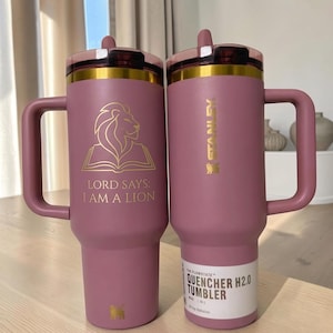 以下が含まれることがあります： ハンドルと蓋付きのダスティローズ色のステンレス製タンブラー2個。片方にはライオンの金色のグラフィックと「LORD SAYS: I AM A LION」の文字。もう片方には「STANLEY」のロゴと「THE FLOWSTATE QUENCHER H2.0 TUMBLER 40 OZ 1.18 L」と書かれたラベル。
