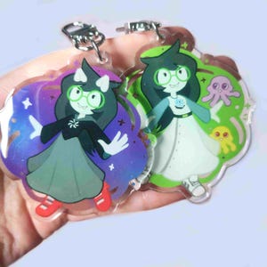 Puede incluir: Dos llaveros acrílicos con personajes de dibujos animados con gafas de montura verde y pelo oscuro. Un llavero tiene un fondo morado y azul, el otro tiene un fondo verde con un pulpo y una criatura amarilla.