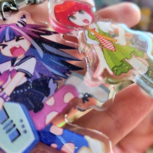 Puede incluir: Dos llaveros de acrílico transparente con personajes de estilo anime y detalles de purpurina. Un personaje tiene el pelo morado y azul en punta, viste ropa oscura y sostiene una guitarra azul claro. El otro tiene el pelo rojo corto y un vestido verde.