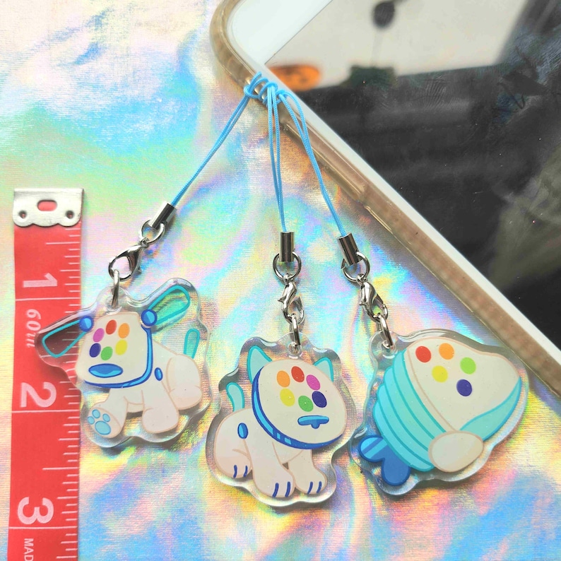 Peut inclure: Trois breloques en acrylique transparent avec des cordons bleus et des fermoirs argent&eacute;s. Les breloques pr&eacute;sentent des motifs de style dessin anim&eacute; d'un chien et d'une forme stylis&eacute;e avec des points color&eacute;s. Un m&egrave;tre ruban rouge et blanc est en arri&egrave;re-plan.