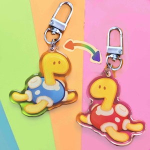May include: Two turtle-shaped keychains with silver clasps. One turtle has a blue shell, the other a red shell. Both have yellow heads, legs, and bellies. A rainbow arrow graphic is between the keychains.