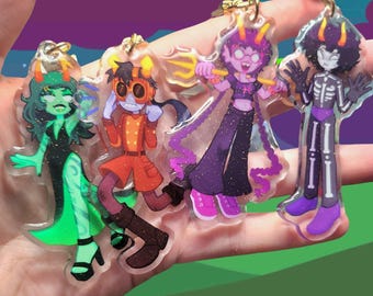 May include: Four clear acrylic keychains featuring stylized cartoon characters with horns. The first is green with a long dress, the second wears an orange coat and goggles, the third is purple with a trident, and the fourth wears a skeleton suit.