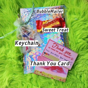 K&ouml;nnte beinhalten: Ein Paket mit einem holografischen Luftpolsterumschlag, einem Schl&uuml;sselanh&auml;nger, einem Starburst-Bonbon und einer Dankeskarte. Der Schl&uuml;sselanh&auml;nger befindet sich in einem kleinen Netzbeutel. Auf der Karte steht "Danke". Der Hintergrund ist hellgr&uuml;n.