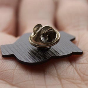 K&ouml;nnte beinhalten: Nahaufnahme einer schwarzen Emaille-Anstecknadel mit goldfarbenem Verschluss. Die Nadel hat eine strukturierte Oberfl&auml;che und eine einzigartige Form. Die Nadel ist wahrscheinlich ein dekoratives Accessoire.