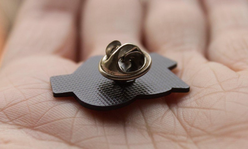 K&ouml;nnte beinhalten: Nahaufnahme einer schwarzen Emaille-Anstecknadel mit goldfarbenem Verschluss. Die Nadel hat eine strukturierte Oberfl&auml;che und eine einzigartige Form. Die Nadel ist wahrscheinlich ein dekoratives Accessoire.