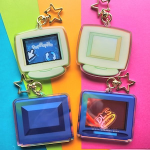 Puede incluir: Cuatro llaveros acrílicos con forma de monitores de ordenador retro. Dos son verdes y dos azules. Los monitores verdes tienen una pantalla con el texto "Popflopita" y el otro tiene una pantalla en blanco. Los monitores azules tienen el texto "DLS LOADING". Cada llavero tiene un dije de estrella dorada.