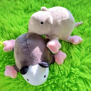 Peut inclure: Deux animaux en peluche, l'un gris clair et l'autre gris, avec des pieds et des oreilles roses, reposent sur une surface verte vif et duveteuse. Les jouets ont des yeux noirs et ressemblent à une souris ou à une créature similaire.