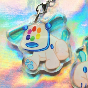 Peut inclure: Un charm en acrylique transparent repr&eacute;sentant un ours de dessin anim&eacute; avec des taches multicolores sur la t&ecirc;te. L'ours a un corps blanc, des accents bleus et un fermoir argent&eacute;. Le charm mesure environ 5 cm de hauteur.