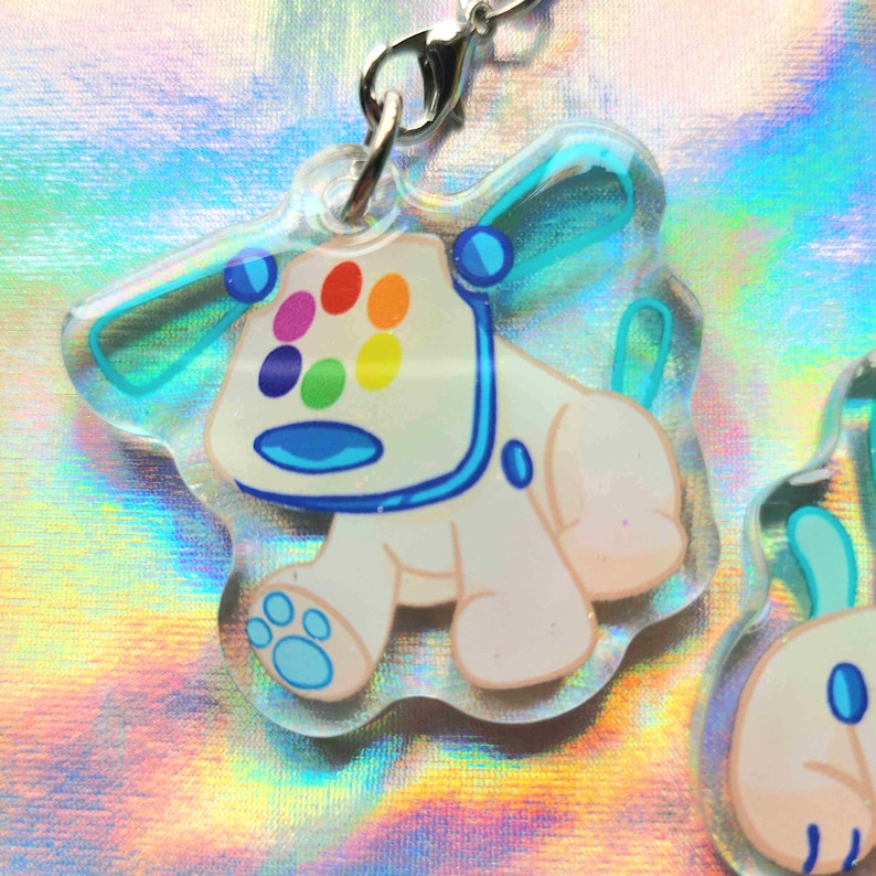Peut inclure: Un charm en acrylique transparent repr&eacute;sentant un ours de dessin anim&eacute; avec des taches multicolores sur la t&ecirc;te. L'ours a un corps blanc, des accents bleus et un fermoir argent&eacute;. Le charm mesure environ 5 cm de hauteur.
