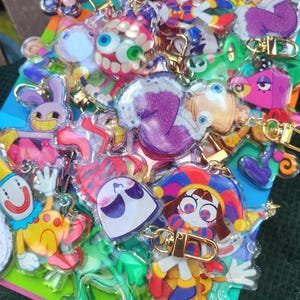 Könnte beinhalten: Eine Sammlung bunter Acryl-Schlüsselanhänger mit verschiedenen Zeichentrickfiguren und Glitzerakzenten. Jeder Anhänger ist an einem goldfarbenen Karabinerverschluss befestigt. Die Motive zeigen Clowns, Narren und verspielte Figuren in leuchtenden Farben wie Violett, Gelb, Rot und Blau.