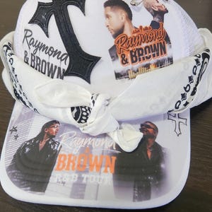 May include: White trucker hat featuring the text "Raymond & Brown" and images of two men. The hat has a black cross and a white mesh back. A white bandana is tied around the hat.