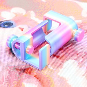 May include: A C-shaped clamp featuring a gradient rainbow finish that transitions between shades of blue, purple, and pink. The device includes two adjustable circular knobs on the sides, designed to tighten or loosen the grip of the clamp mechanism.