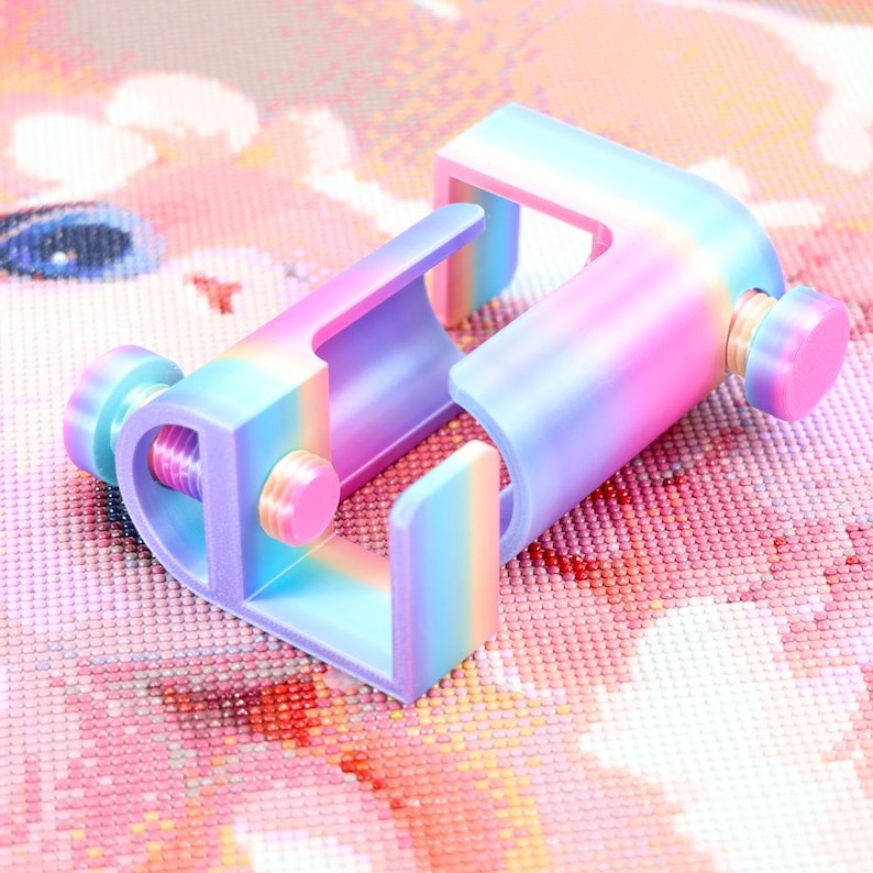 May include: A C-shaped clamp featuring a gradient rainbow finish that transitions between shades of blue, purple, and pink. The device includes two adjustable circular knobs on the sides, designed to tighten or loosen the grip of the clamp mechanism.