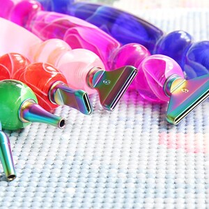 May include: A collection of colorful diamond painting pens. The pens have a variety of colors, including red, green, blue, pink, and purple. Each pen has a metallic tip and a rounded, clear body. The pens are arranged on a light blue, textured surface.