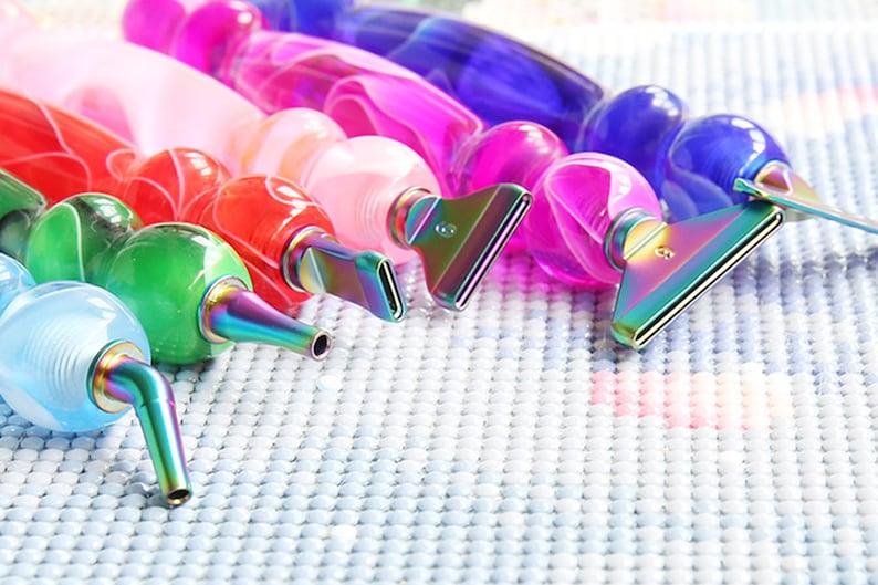 May include: A collection of colorful diamond painting pens. The pens have a variety of colors, including red, green, blue, pink, and purple. Each pen has a metallic tip and a rounded, clear body. The pens are arranged on a light blue, textured surface.