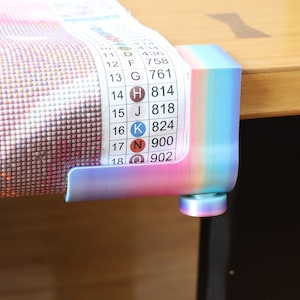 May include: A rainbow-coloured gradient plastic clamp holding a diamond painting canvas. The canvas features a printed legend with numbers 10 through 18, corresponding letters, and colour-coded symbols. The clamp has a circular tightening knob.