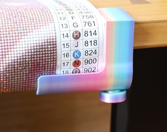 Puede incluir: Una abrazadera de plástico con degradado de arcoíris que sujeta un lienzo de diamond painting. El lienzo presenta una leyenda impresa con los números del 10 al 18, letras correspondientes y símbolos codificados por colores. Tiene una perilla.