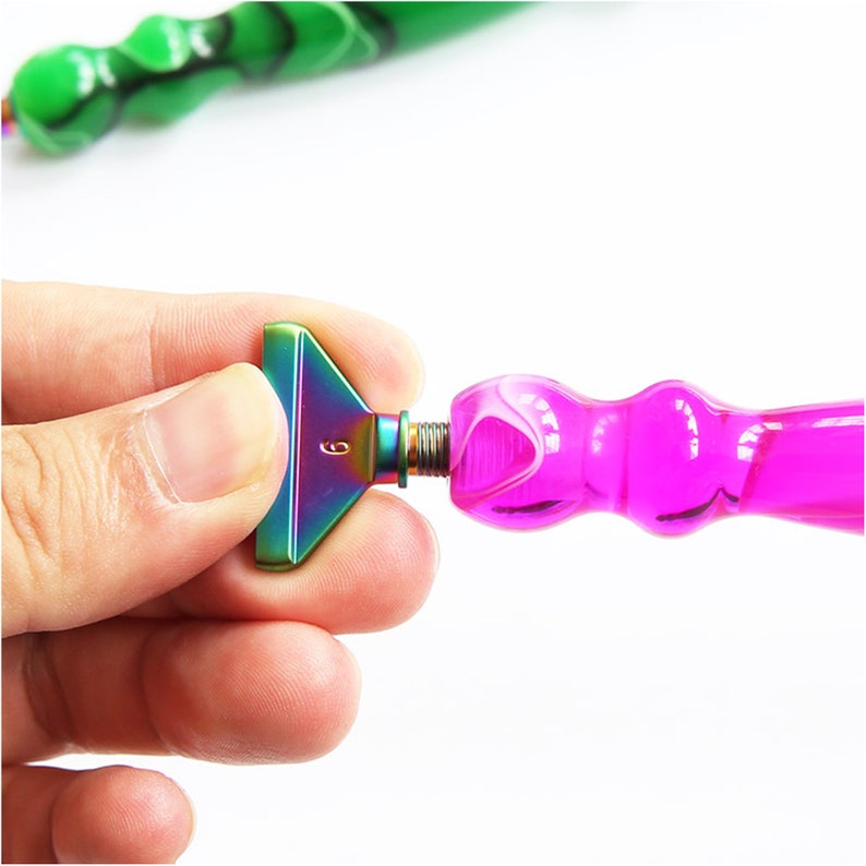 May include: A close-up of a diamond painting pen with a rainbow-colored metal tip and a pink handle. The tip is shaped like a small, flat triangle with the number 6 engraved on it. The pen is designed for precision and detail work.