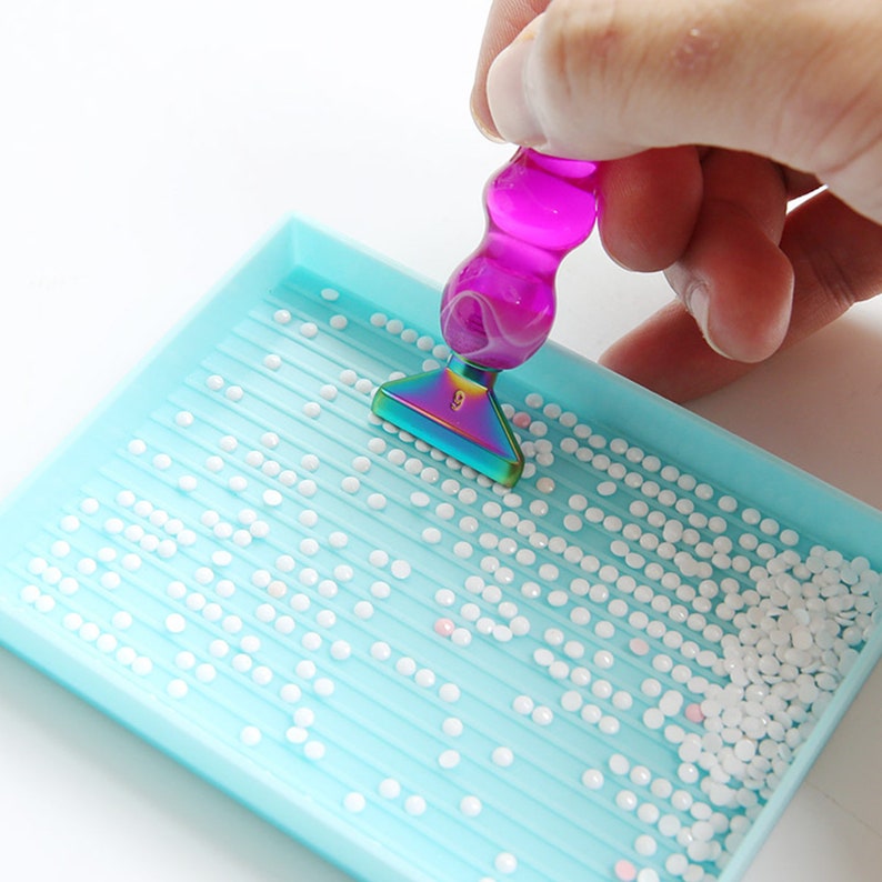 May include: A close-up of a diamond painting kit. A light blue tray is filled with white, round rhinestones. A pink and iridescent-tipped pen is positioned above the tray. The pen has a rainbow-colored tip with the number 9.