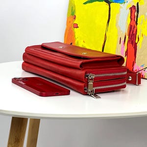 May include: A rectangular red leather wallet featuring a fold-over flap with two silver-toned snap buttons and two separate zippered compartments with silver metal zippers. A matching red smartphone is positioned flat next to the wallet.