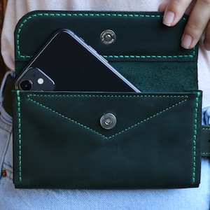 May include: A dark green leather phone case with a green stitching detail. The case is attached to a belt with a metal snap closure.
