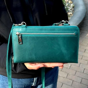 May include: A rectangular teal leather crossbody bag featuring a horizontal zippered pocket on the front, a silver-toned metal zipper pull, and a matching thin leather shoulder strap. The bag has visible stitching along the top edge and sides.