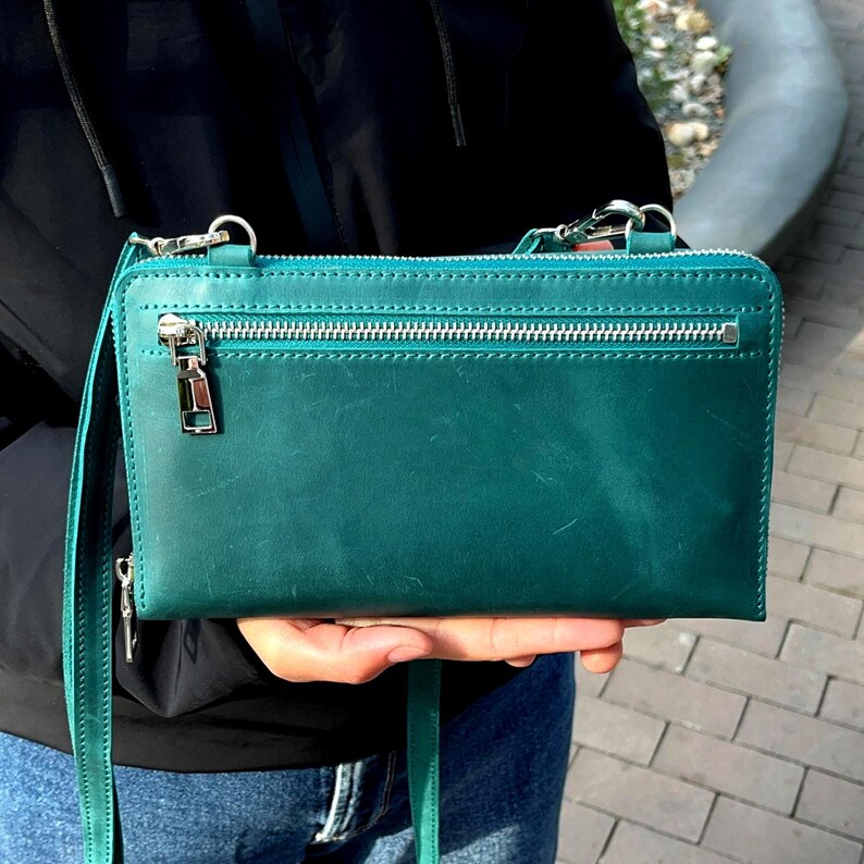 May include: A rectangular teal leather crossbody bag featuring a horizontal zippered pocket on the front, a silver-toned metal zipper pull, and a matching thin leather shoulder strap. The bag has visible stitching along the top edge and sides.