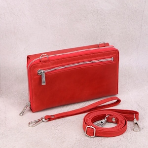 May include: A rectangular red leather wallet featuring a top zipper closure, a secondary front zipper pocket, and silver-toned metal hardware. The wallet includes a detachable red wrist strap and a longer adjustable red shoulder strap with clips.
