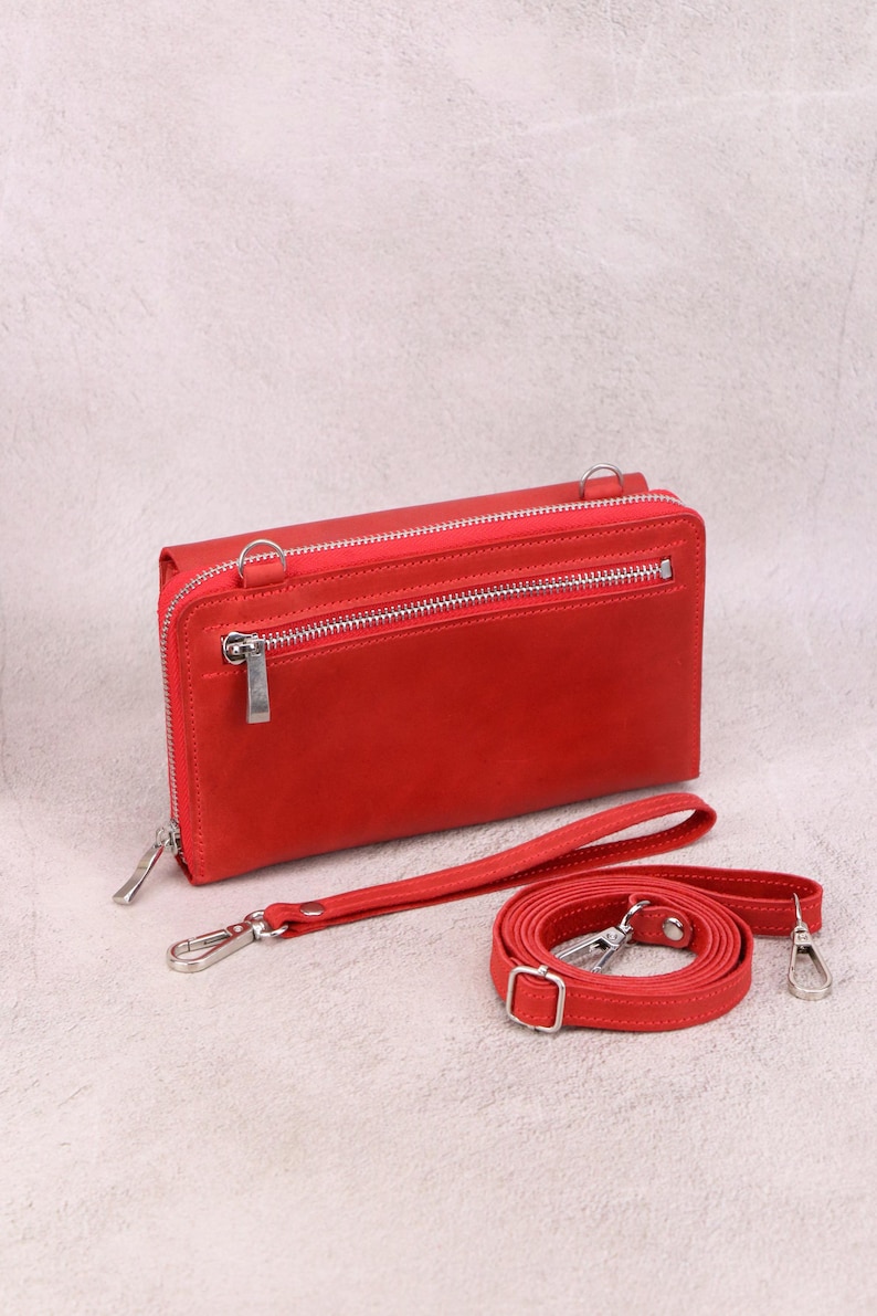 May include: A rectangular red leather wallet featuring a top zipper closure, a secondary front zipper pocket, and silver-toned metal hardware. The wallet includes a detachable red wrist strap and a longer adjustable red shoulder strap with clips.