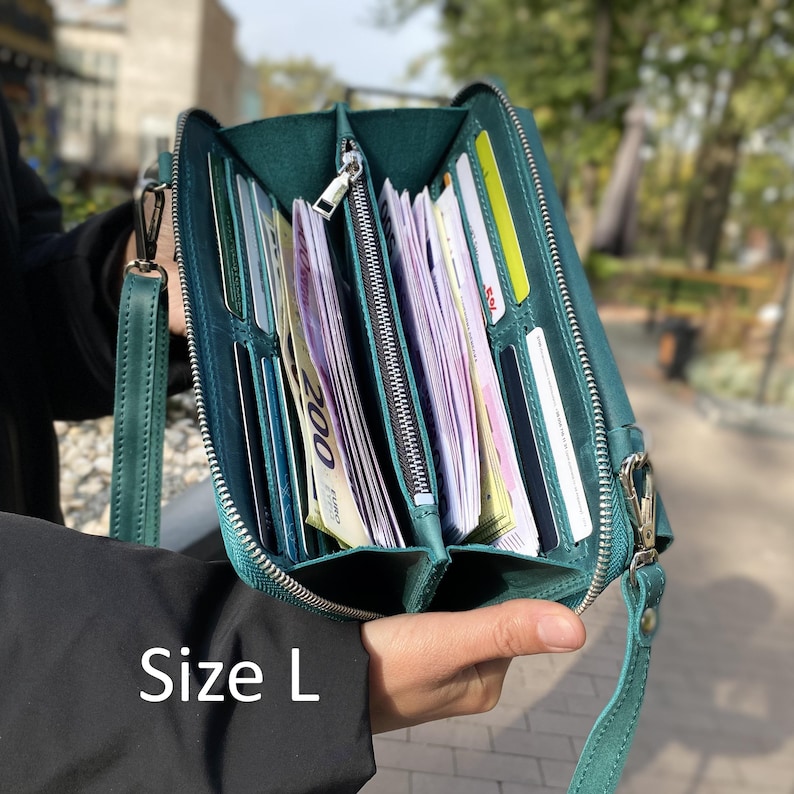 May include: A teal leather wallet with a zipper closure, featuring multiple interior card slots and compartments filled with paper currency. The wallet includes a detachable wrist strap and a metal clasp. The text Size L is visible.