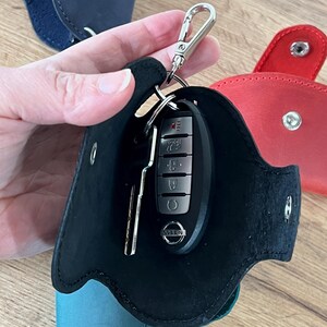 May include: A black leather key fob holder with a silver clasp, holding a car key. The holder is open, revealing the key and a key. Other key fob holders in blue, red, and teal are visible in the background.