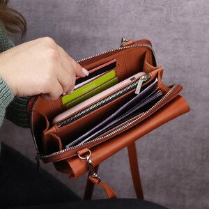 May include: A tan leather crossbody bag with a zippered main compartment, featuring multiple interior slots for credit cards, a central zippered pocket, and an open section currently holding a smartphone and several folded paper documents.