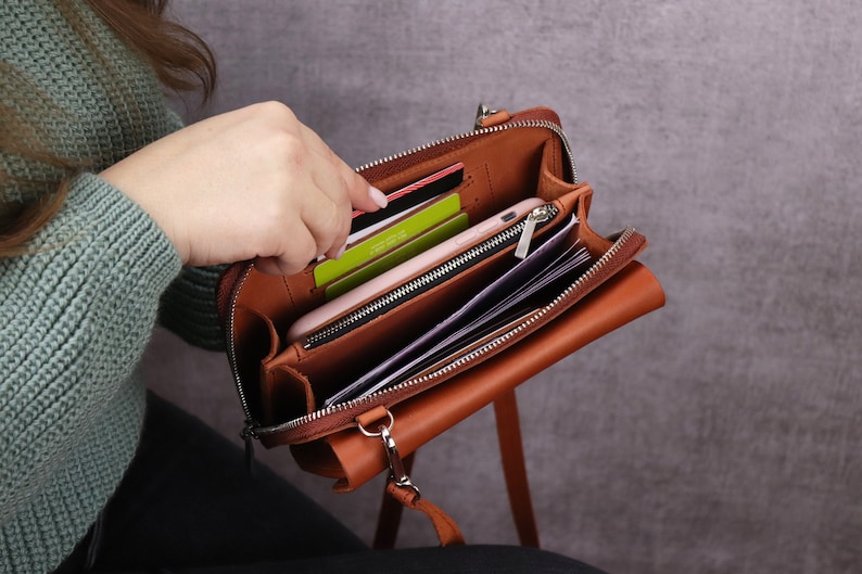 May include: A tan leather crossbody bag with a zippered main compartment, featuring multiple interior slots for credit cards, a central zippered pocket, and an open section currently holding a smartphone and several folded paper documents.
