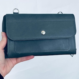 May include: A dark gray rectangular leather wallet featuring a front flap with a single silver-toned metal snap button closure, visible stitching along the edges, and two silver metal D-rings attached to the top for a shoulder strap.