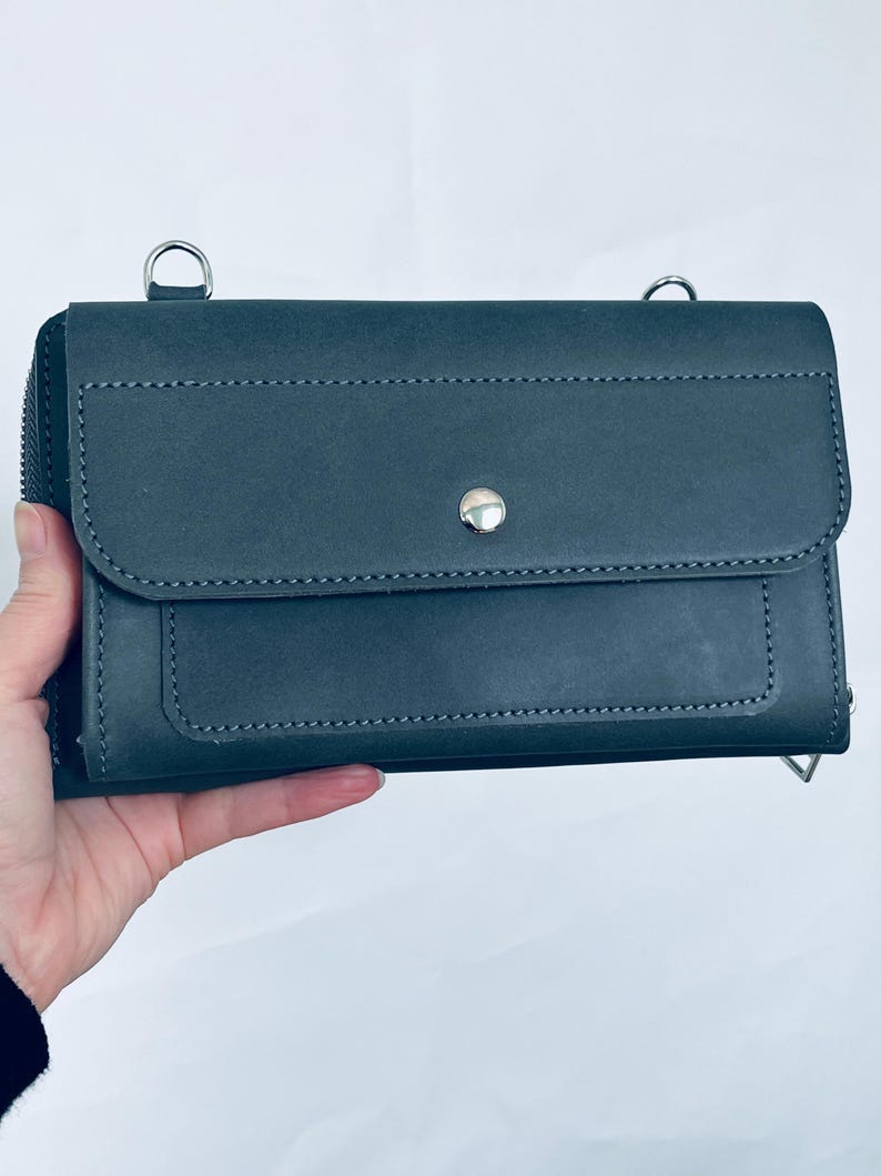 May include: A dark gray rectangular leather wallet featuring a front flap with a single silver-toned metal snap button closure, visible stitching along the edges, and two silver metal D-rings attached to the top for a shoulder strap.